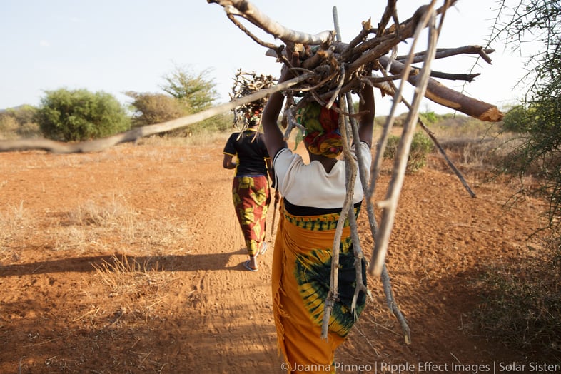 Two women on a dirt path, carrying bundles of sticks.